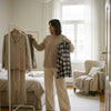 Volwassen vrouw vergelijkt twee pyjama’s in een rustige slaapkamer, met zachte ochtendlichtinval en focus op stof, pasvorm en comfort.