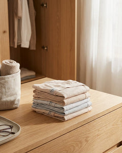 Neatly folded Sloggi Basic cotton underwear in soft neutral tones on a bright bedroom dresser, styled to suggest everyday comfort and trusted basics.